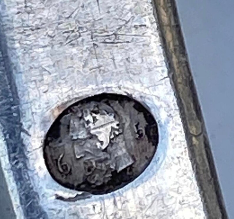 May include: Close-up of a silver object with a circular mark. The mark is a profile of a person's head with the number "16" visible.