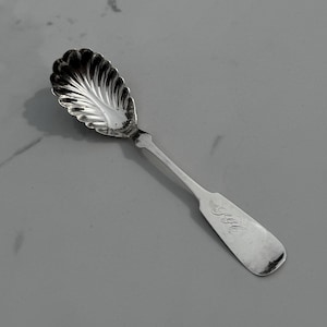 May include: A silver spoon with a shell-shaped bowl and a long, rectangular handle. The handle is engraved with the initials "S.H.". The spoon is made of a shiny, reflective metal.
