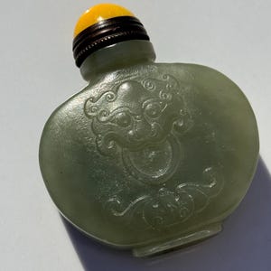 May include: A pale green, translucent snuff bottle featuring a carved relief of a stylized creature face on the front. The bottle is topped with a dark brown, ribbed metal collar and a smooth, rounded, bright yellow stopper.
