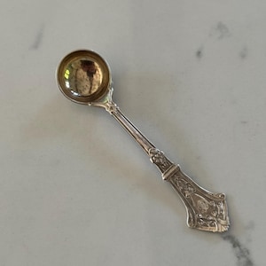 Puede incluir: Una cuchara de plata con un cuenco pequeño y redondo y un mango adornado. El mango presenta diseños detallados de flores y volutas. La cuchara es probablemente vintage o antigua, y está diseñada para servir o comer.