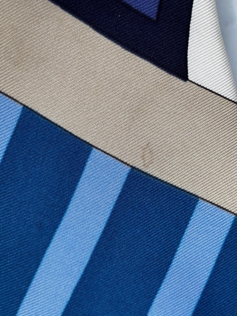Puede incluir: Primer plano de un pa&ntilde;uelo de seda con un dise&ntilde;o geom&eacute;trico. El pa&ntilde;uelo presenta una combinaci&oacute;n de colores azul, azul claro, beige y azul marino. Rayas diagonales y formas rectangulares crean un patr&oacute;n moderno.