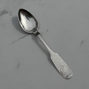 May include: A silver spoon with an oval bowl and a long, slender handle. The handle has a rectangular end with an engraved design. The spoon is made of a shiny, reflective metal.