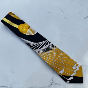 Puede incluir: Un pa&ntilde;uelo de seda negro y dorado con un dise&ntilde;o tem&aacute;tico de golf. El pa&ntilde;uelo presenta un patr&oacute;n de pelotas de golf, palos de golf y l&iacute;neas geom&eacute;tricas. El pa&ntilde;uelo mide aproximadamente 7.6 cm de ancho y 101.6 cm de largo.