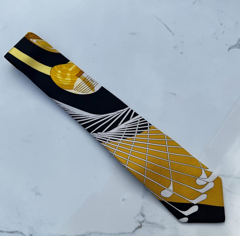 Puede incluir: Un pa&ntilde;uelo de seda negro y dorado con un dise&ntilde;o tem&aacute;tico de golf. El pa&ntilde;uelo presenta un patr&oacute;n de pelotas de golf, palos de golf y l&iacute;neas geom&eacute;tricas. El pa&ntilde;uelo mide aproximadamente 7.6 cm de ancho y 101.6 cm de largo.