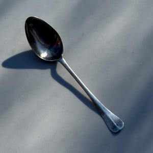 May include: A silver spoon with a rounded bowl and a long, slender handle. The spoon is positioned diagonally, casting a shadow on the gray surface. The spoon's surface reflects light, highlighting its polished finish. A simple, elegant utensil.