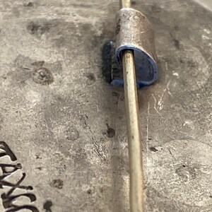 May include: Close-up of the back of a silver pin with a metal pin back. The pin has the word "TAVIK" engraved on the back.