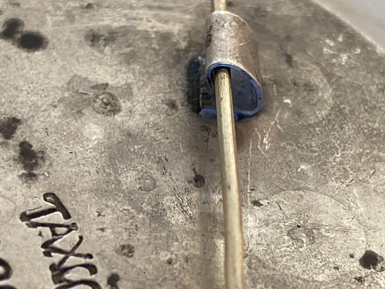 May include: Close-up of the back of a silver pin with a metal pin back. The pin has the word "TAVIK" engraved on the back.