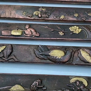 K&ouml;nnte beinhalten: F&uuml;nf dekorative Metallplatten mit aufwendigen Designs. Jede Platte zeigt eine andere Szene mit Figuren, V&ouml;geln und floralen Motiven. Die Platten sind aus dunklem Metall mit goldenen Akzenten.