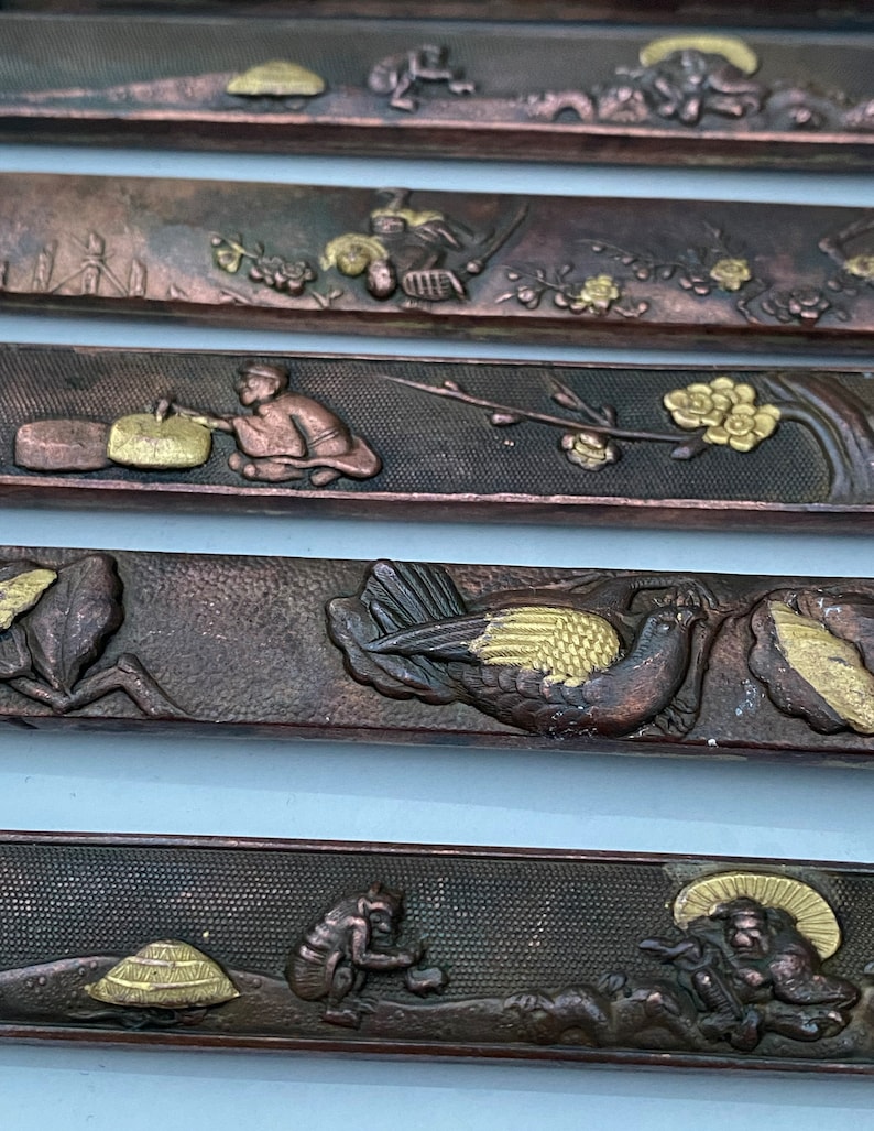 K&ouml;nnte beinhalten: F&uuml;nf dekorative Metallplatten mit aufwendigen Designs. Jede Platte zeigt eine andere Szene mit Figuren, V&ouml;geln und floralen Motiven. Die Platten sind aus dunklem Metall mit goldenen Akzenten.