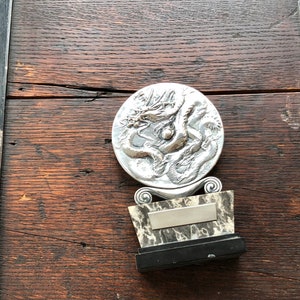 May include: A silver dragon sculpture on a black and white marble base with a silver plaque. The sculpture is mounted on a dark wood surface.