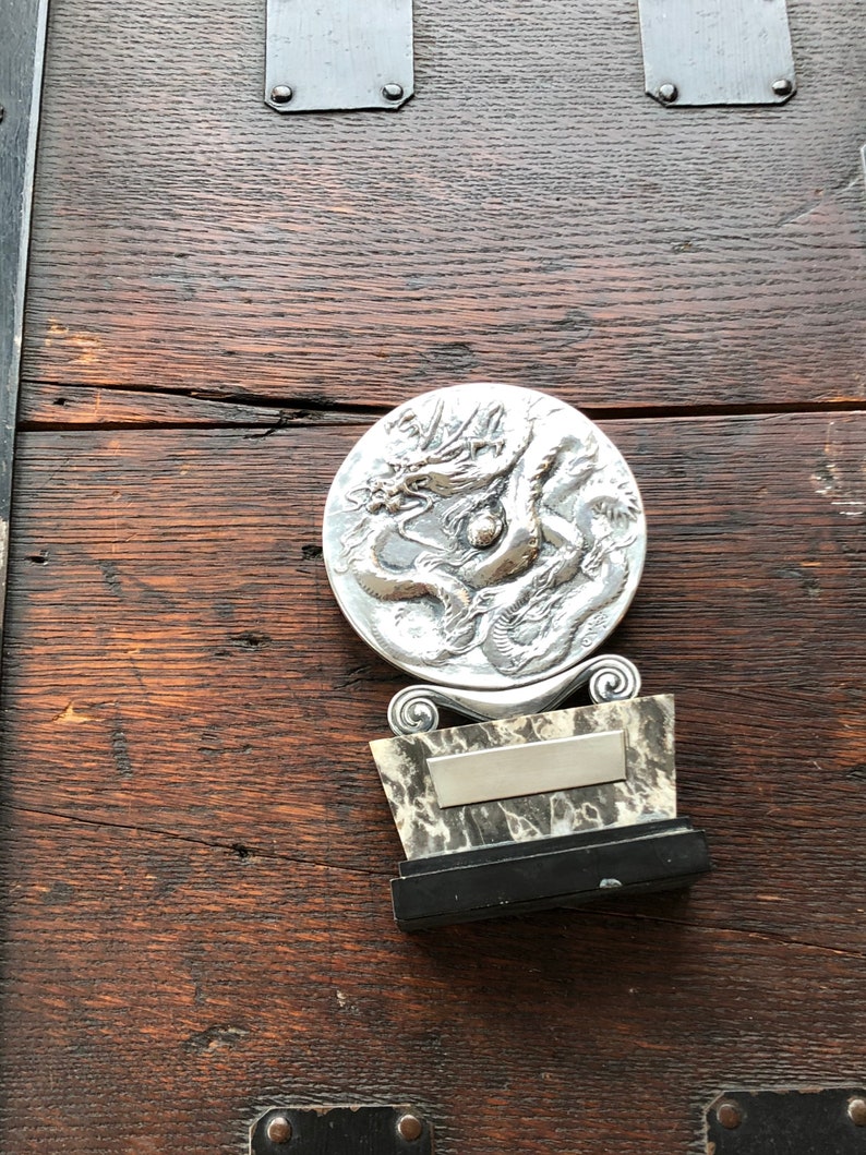 May include: A silver dragon sculpture on a black and white marble base with a silver plaque. The sculpture is mounted on a dark wood surface.