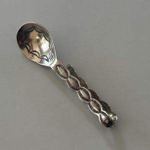 Peut inclure: Une cuillère en argent avec un bol ovale et un manche orné d'un motif ovale complexe et répétitif. Le bol de la cuillère présente un motif en forme d'éclat d'étoile. La cuillère est probablement en argent sterling.