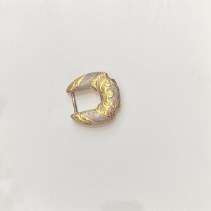 May include: A gold-toned metal buckle with a decorative design. The buckle has a silver-toned finish and a floral pattern.