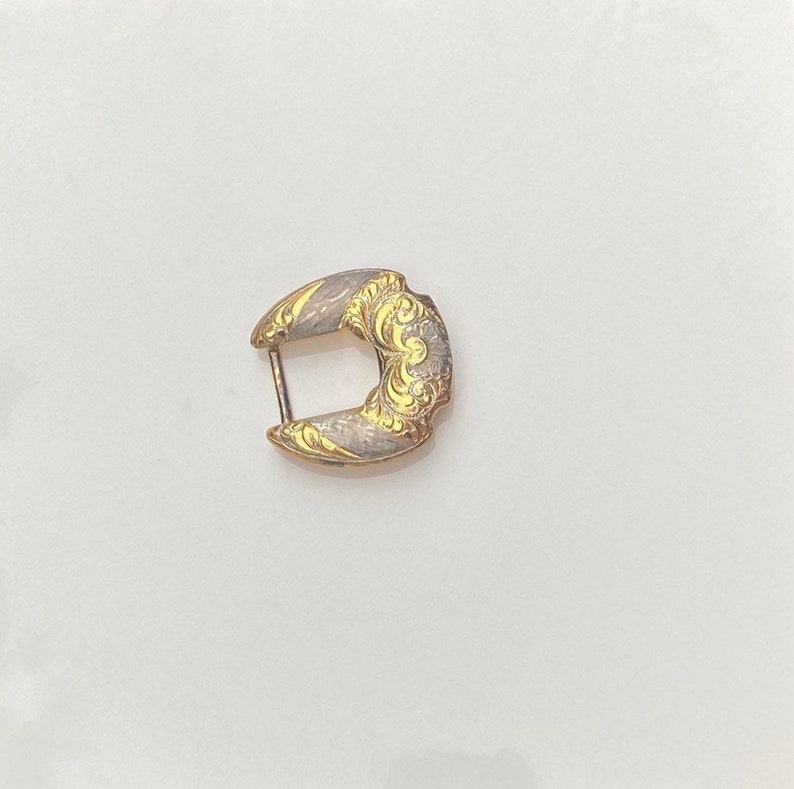 May include: A gold-toned metal buckle with a decorative design. The buckle has a silver-toned finish and a floral pattern.