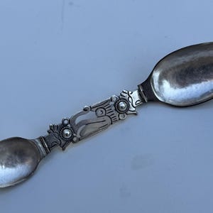 Könnte beinhalten: Ein silberner Löffel mit einem verzierten Griff. Der Löffel hat zwei ovale Schalen, eine an jedem Ende, und einen dekorativen Griff mit floralen und scrollartigen Designs. Der Löffel ist wahrscheinlich aus Sterlingsilber.