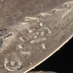 May include: Close-up of a silver-toned metal object with a circular shape and a stamped inscription. The inscription is difficult to read, but it appears to be a series of letters or numbers.