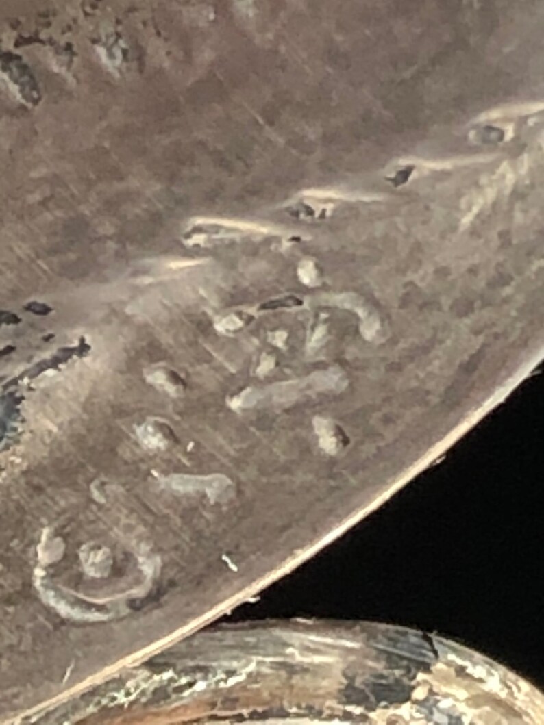 May include: Close-up of a silver-toned metal object with a circular shape and a stamped inscription. The inscription is difficult to read, but it appears to be a series of letters or numbers.