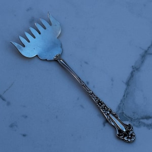 以下が含まれることがあります： 銀色のサービングフォーク。柄には花の装飾が施されています。フォークは、複数の歯が付いた、幅広の扇形のヘッドを備えています。柄は細く、繊細な花のディテールが特徴です。
