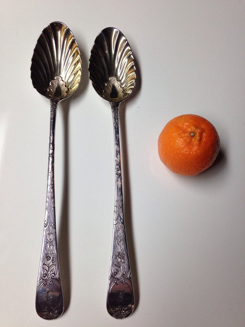 May include: Two ornate silver spoons with shell-shaped bowls and detailed floral patterns on the handles. An orange tangerine sits to the side. The spoons are likely for serving or display.