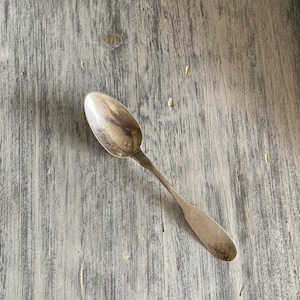 May include: A silver teaspoon with a rounded bowl and a long, slender handle. The handle is engraved with the word "Marguerite".