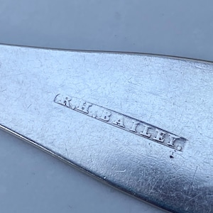 May include: Close-up of a silver utensil with the inscription "R.H. BAILEY." The inscription is engraved on the back of the utensil.