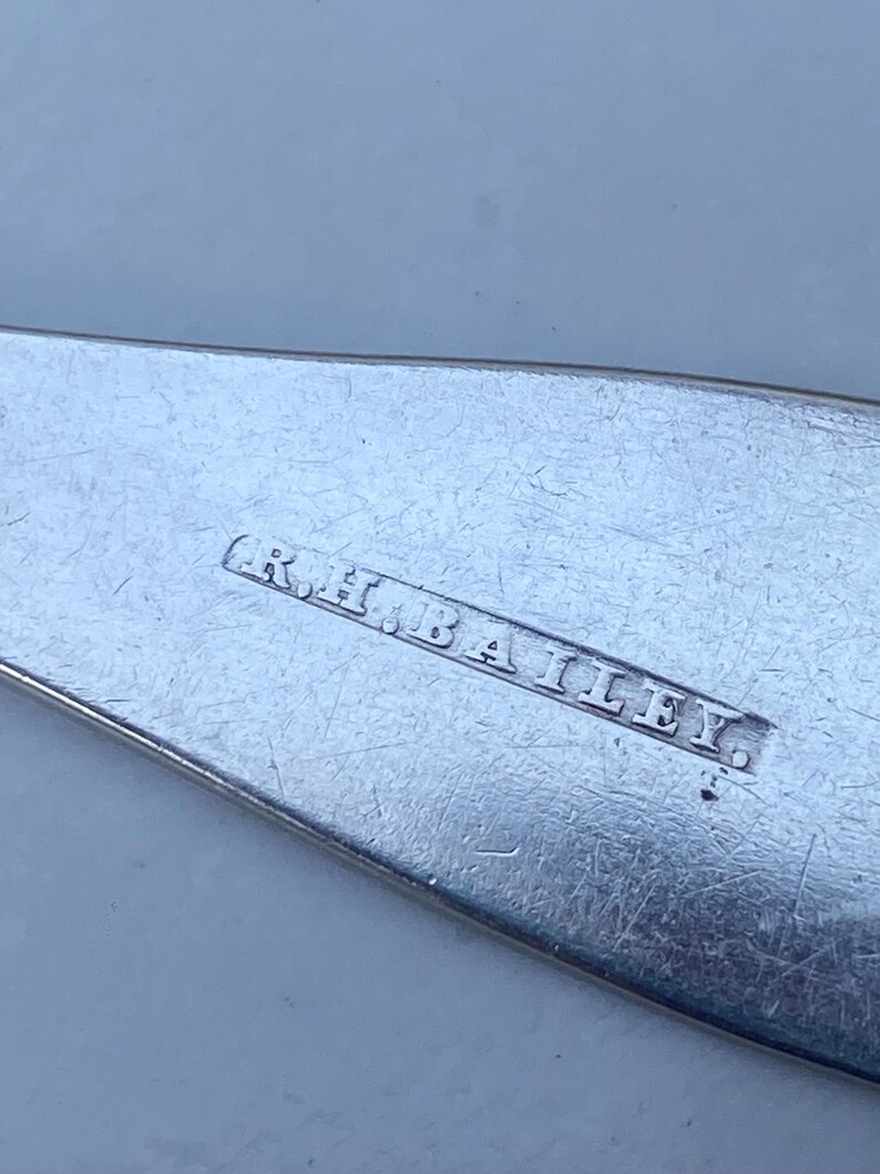 May include: Close-up of a silver utensil with the inscription "R.H. BAILEY." The inscription is engraved on the back of the utensil.