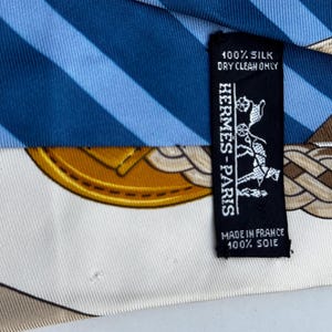 Puede incluir: Un pa&ntilde;uelo de seda con rayas azules y blancas, un dise&ntilde;o de medall&oacute;n dorado y un patr&oacute;n de cuerda marr&oacute;n. Una etiqueta negra dice "HERMES-PARIS" y "100% Silk, Dry Clean Only, Made in France, 100% Soie."