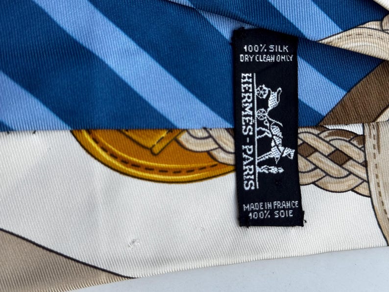 Puede incluir: Un pa&ntilde;uelo de seda con rayas azules y blancas, un dise&ntilde;o de medall&oacute;n dorado y un patr&oacute;n de cuerda marr&oacute;n. Una etiqueta negra dice "HERMES-PARIS" y "100% Silk, Dry Clean Only, Made in France, 100% Soie."