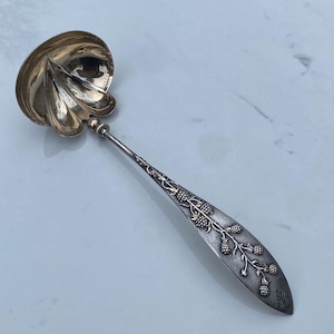 May include: A silver serving spoon with a decorative handle featuring a vine of berries. The bowl of the spoon is shaped like a leaf and is polished to a shine.