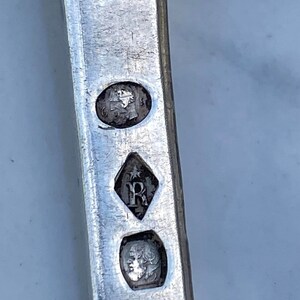 May include: Close-up of a silver object with three hallmarks. The top hallmark is a circular stamp with a profile of a person's head. The middle hallmark is a diamond-shaped stamp with a star and the letter "R". The bottom hallmark is a circular stamp with a profile of a person's head.