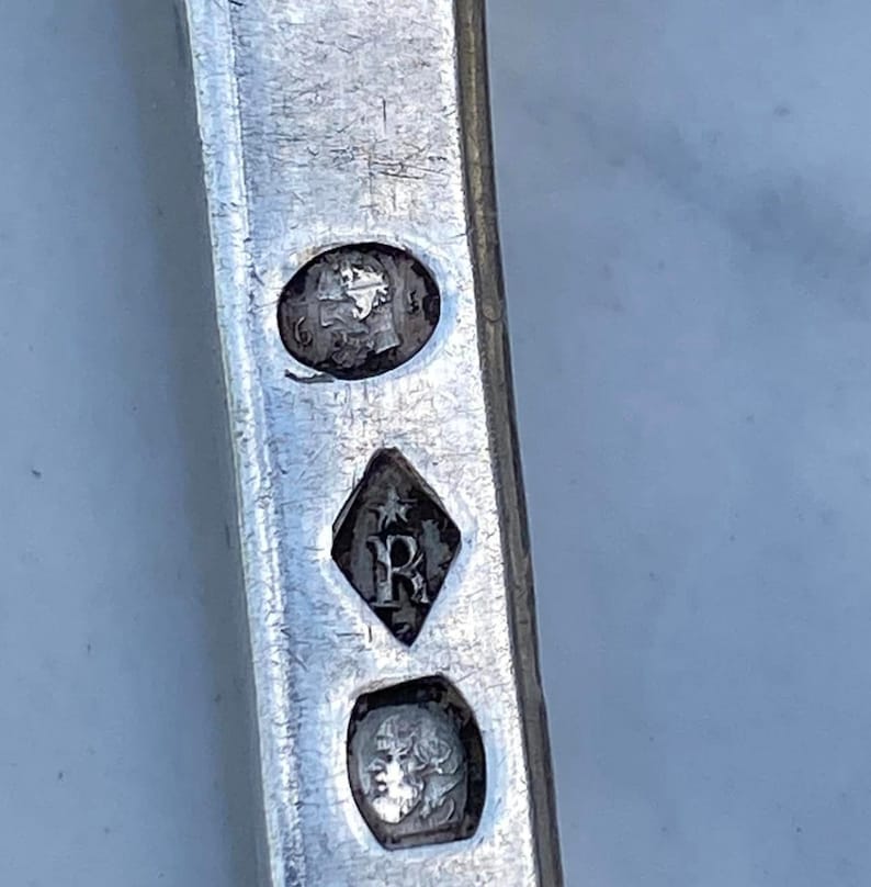 May include: Close-up of a silver object with three hallmarks. The top hallmark is a circular stamp with a profile of a person's head. The middle hallmark is a diamond-shaped stamp with a star and the letter "R". The bottom hallmark is a circular stamp with a profile of a person's head.