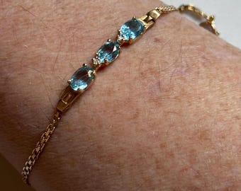May include: A gold bracelet featuring three oval, light blue gemstones. The bracelet has a delicate chain and is adorned with small accent stones. The bracelet is shown on a person's wrist.