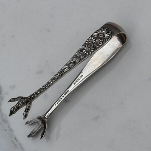 May include: A pair of silver sugar tongs featuring a floral embossed pattern on the handles and claw-shaped ends. The metal is stamped with the text KIRK & SON and STERLING. The tongs have a polished finish and a curved, spring-loaded design.