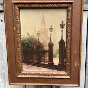 Puede incluir: Una fotografía antigua enmarcada de una escena callejera en Nueva Orleans, Luisiana. La imagen presenta una iglesia con una alta aguja, farolas y una puerta de hierro forjado. La fotografía está en tonos sepia y tiene un aspecto descolorido y antiguo.