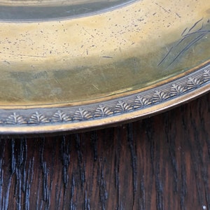 Puede incluir: Primer plano del borde de un plato redondo de color dorado con un borde decorativo. El plato est&aacute; apoyado sobre una superficie de madera oscura.