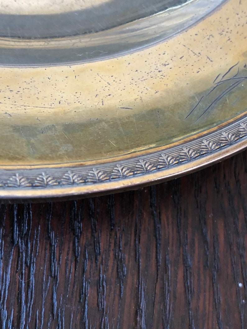 Puede incluir: Primer plano del borde de un plato redondo de color dorado con un borde decorativo. El plato est&aacute; apoyado sobre una superficie de madera oscura.