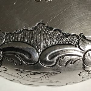 以下が含まれることがあります： 装飾的なデザインと上部に刻印された「U」の文字を持つ銀製の物体のクローズアップ。