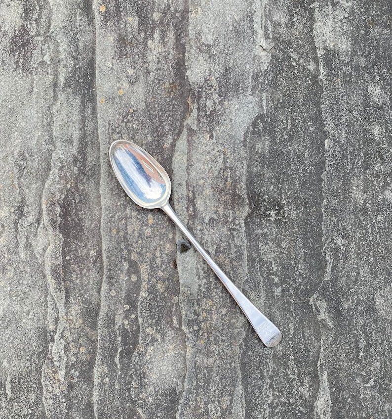May include: A silver teaspoon with a rounded bowl and a long, slender handle. The spoon is lying on a textured, gray surface.