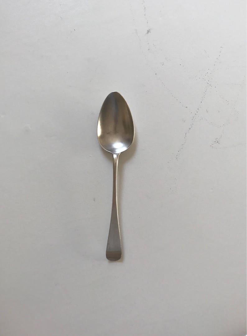 Może przedstawiać: Srebrna łyżka z owalną miseczką i długim, smukłym trzonkiem. Uchwyt ma płaski, prostokątny koniec. Łyżka jest umieszczona na białym tle, odbijając światło.