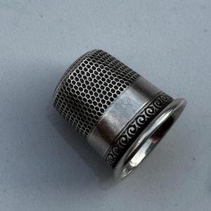 Op de afbeelding: Een zilveren vingerhoed met een honingraatpatroon aan de bovenkant en een decoratief wervelontwerp rond de basis. Dit naai-instrument is ontworpen om de vinger te beschermen bij het doorsteken van een naald door de stof.