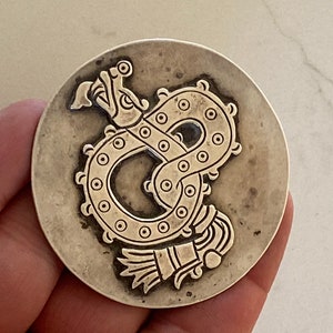 May include: Silver pendant featuring a detailed embossed design of a serpent with a feathered headdress. The pendant is circular and has a slightly textured surface.
