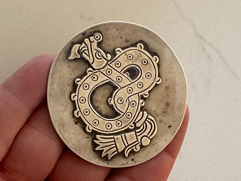 May include: Silver pendant featuring a detailed embossed design of a serpent with a feathered headdress. The pendant is circular and has a slightly textured surface.