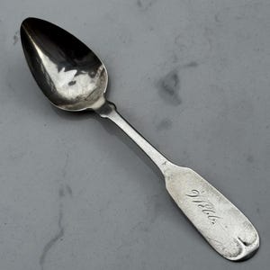 May include: A silver spoon with a rounded bowl and a long, slender handle. The handle has a rectangular end with the engraved word "Webb." The spoon is likely vintage or antique.
