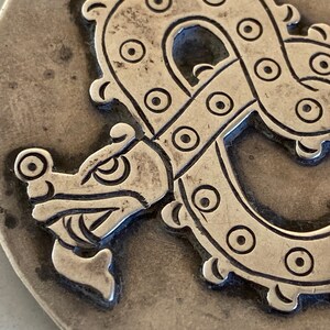 May include: A silver-toned metal pendant featuring a detailed embossed design of a stylized dragon with a circular pattern.