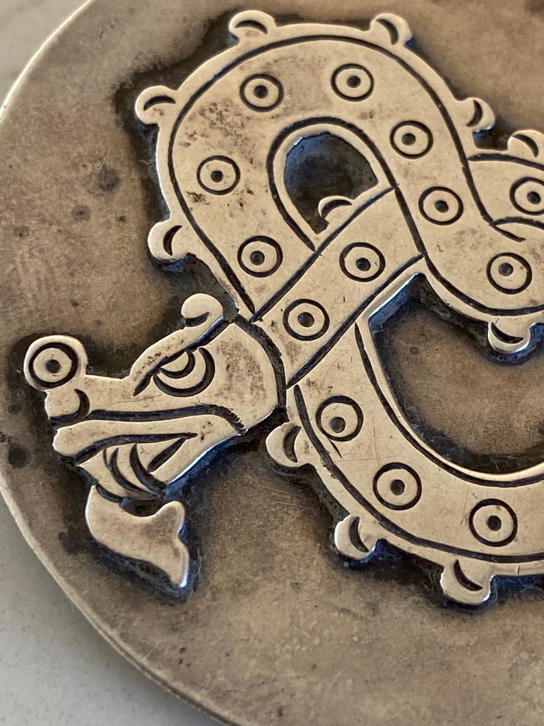 May include: A silver-toned metal pendant featuring a detailed embossed design of a stylized dragon with a circular pattern.