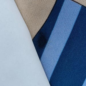 Puede incluir: Primer plano de una corbata de seda doblada con un patr&oacute;n de rayas en tonos azules. Un trozo de papel blanco cubre parcialmente la corbata, revelando una secci&oacute;n de tela beige. La tela tiene un brillo sutil.