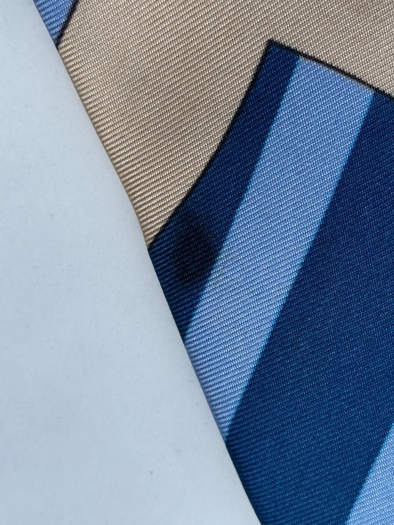 Puede incluir: Primer plano de una corbata de seda doblada con un patr&oacute;n de rayas en tonos azules. Un trozo de papel blanco cubre parcialmente la corbata, revelando una secci&oacute;n de tela beige. La tela tiene un brillo sutil.