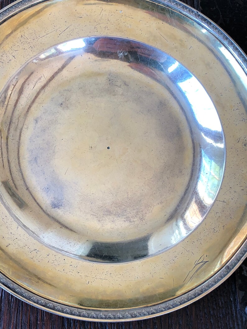 Puede incluir: Un plato de plata con un borde decorativo y una peque&ntilde;a marca en el centro.