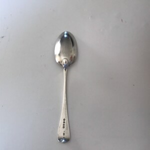 Może przedstawiać: Srebrna łyżka z zaokrągloną miseczką i długim, smukłym trzonkiem. Uchwyt ma oznaczenia w pobliżu końca. Łyżka jest odblaskowa, a powierzchnia, na kt&oacute;rej spoczywa, jest jasnoszara. Łyżka jest przedmiotem vintage.