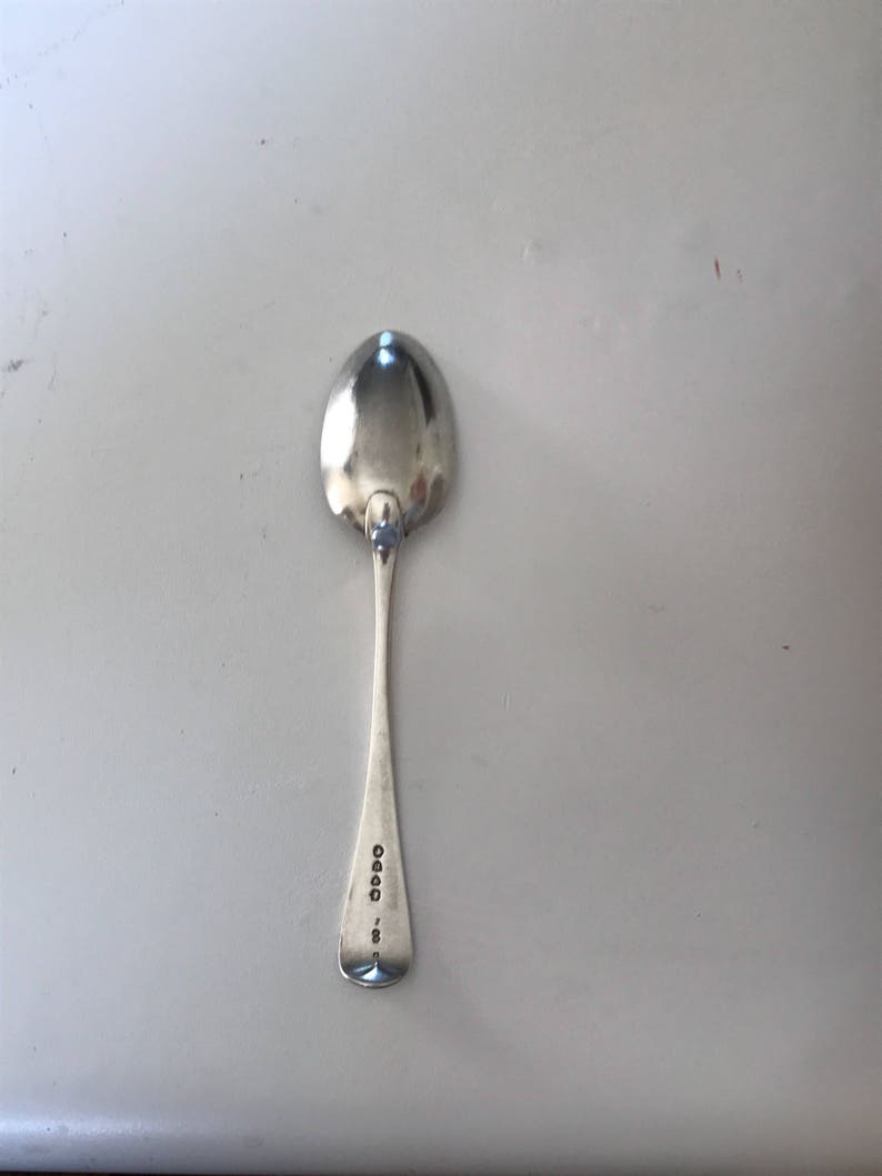 Może przedstawiać: Srebrna łyżka z zaokrągloną miseczką i długim, smukłym trzonkiem. Uchwyt ma oznaczenia w pobliżu końca. Łyżka jest odblaskowa, a powierzchnia, na kt&oacute;rej spoczywa, jest jasnoszara. Łyżka jest przedmiotem vintage.